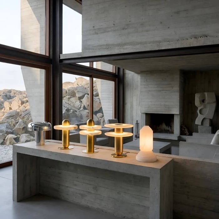 Modern concrete interior with floor-to-ceiling windows overlooking rocky terrain, featuring a row of sculptural table lamps in brass, chrome, and frosted glass displayed on a бетон console near a fireplace.