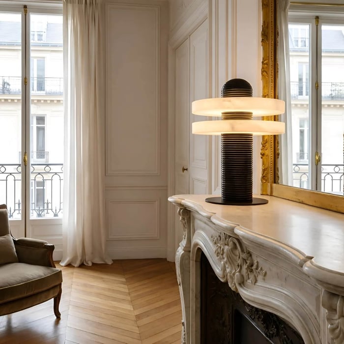 Elegant Parisian-style living room with ornate white fireplace mantel and gold mirror, featuring a ribbed black alabaster table lamps with a double-ring illuminated shade placed on the mantel near tall French windows.
