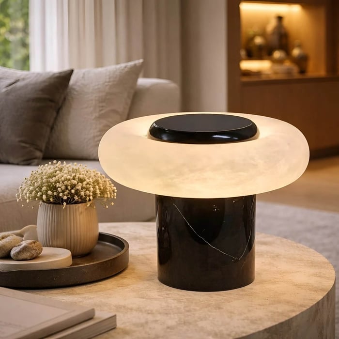 Close-up of a modern marble-based table lamp with a thick circular illuminated shade on a stone side table, styled with decorative objects in a warm neutral living space.