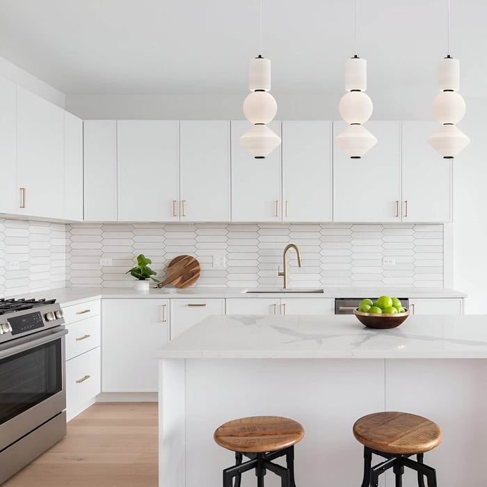Bright white kitchen with gold hardware, wooden stools, and three modern white pendant lights above the island.