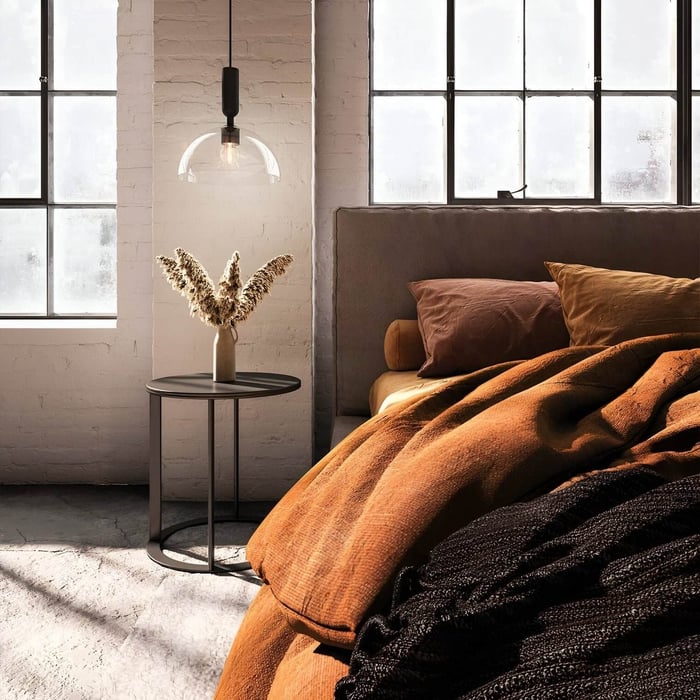Minimalist pendant light with clear glass shade hanging by a bed in an industrial-style room.