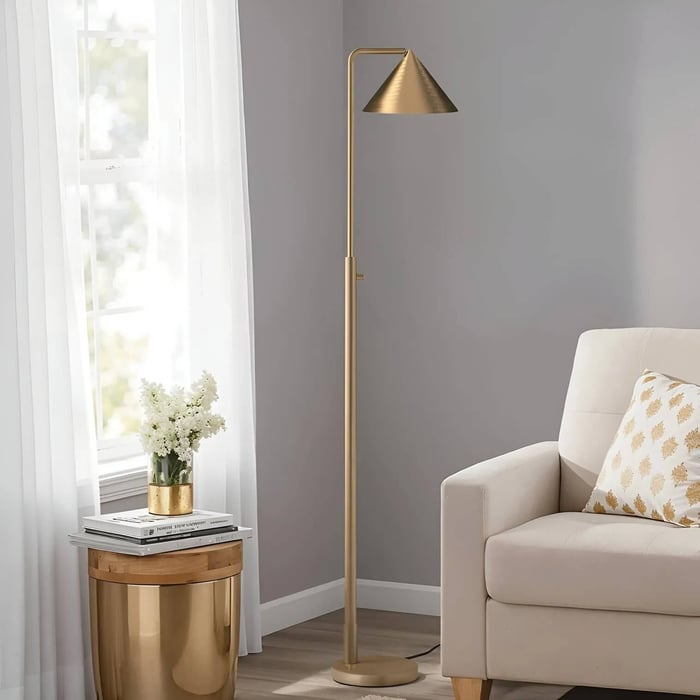 Tall brass industrial floor lamp near a window, beside a beige sofa and gold side table in a modern living room.