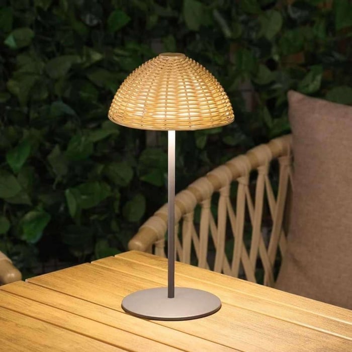 Rattan-style outdoor table lamp glowing on a wooden patio table, next to a woven chair with cushions and leafy greenery in the background.