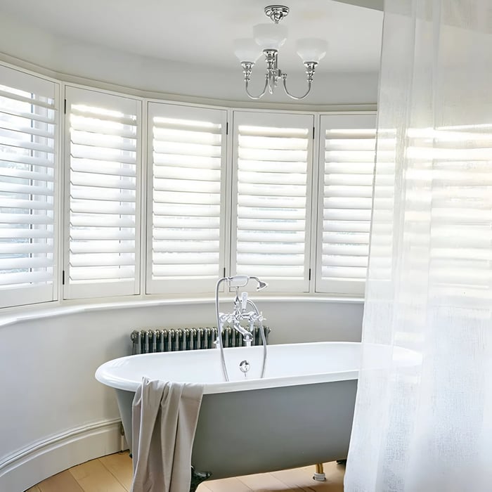 The image shows a bright bathroom with a freestanding white and gray clawfoot bathtub positioned in front of a curved set of white plantation shutters, allowing in soft, diffused light. A chrome faucet fixture and a small towel draped over the tub's edge add a touch of luxury. A three-light chandelier with frosted glass shades hangs from the ceiling, contributing to the serene and elegant atmosphere. Sheer curtains to the side add a soft, airy feel to the room.