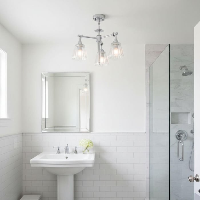 Bright white bathroom with a pedestal sink, framed mirror, chrome three-light ceiling fixture, and a glass walk-in shower on the right.