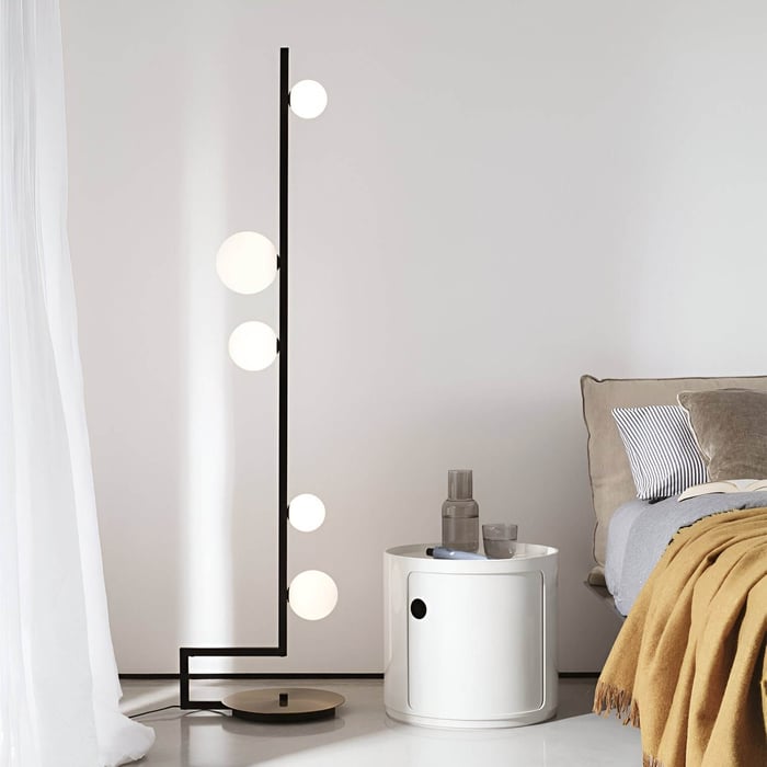 Minimalist bedroom corner with a black vertical floor lamp holding spherical bulbs, next to a white round nightstand.
