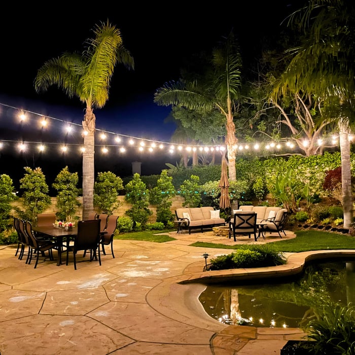 Warm outdoor string lights stretched across a landscaped patio with palm trees, dining table, and lounge seating at night.