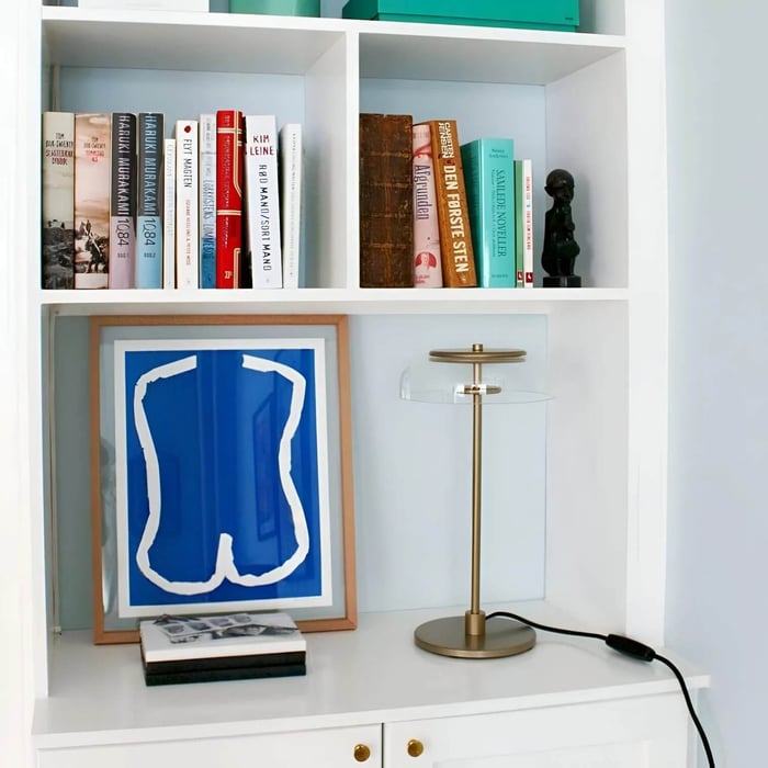 Slim brass table lamp with a clear circular shade on a white bookshelf beside books and framed art.