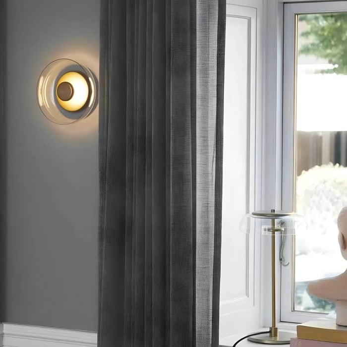 Round glass wall light with gold center beside gray curtains and a window in a modern interior.