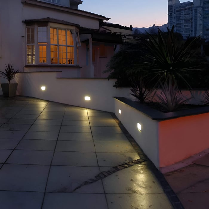 House patio at dusk with square recessed wall lights guiding a paved walkway alongside raised planters and exterior walls.