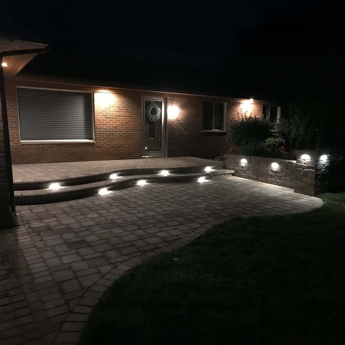 Night view of a brick patio with wall and step lights casting a soft glow on pavers and landscaped borders.