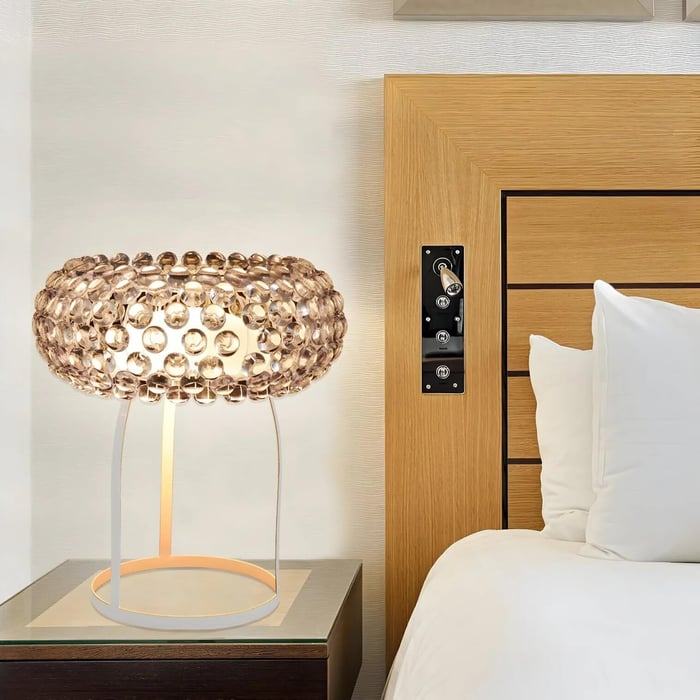 Table lamp with a sparkling beaded shade on a bedside table next to a wooden headboard and white bedding.