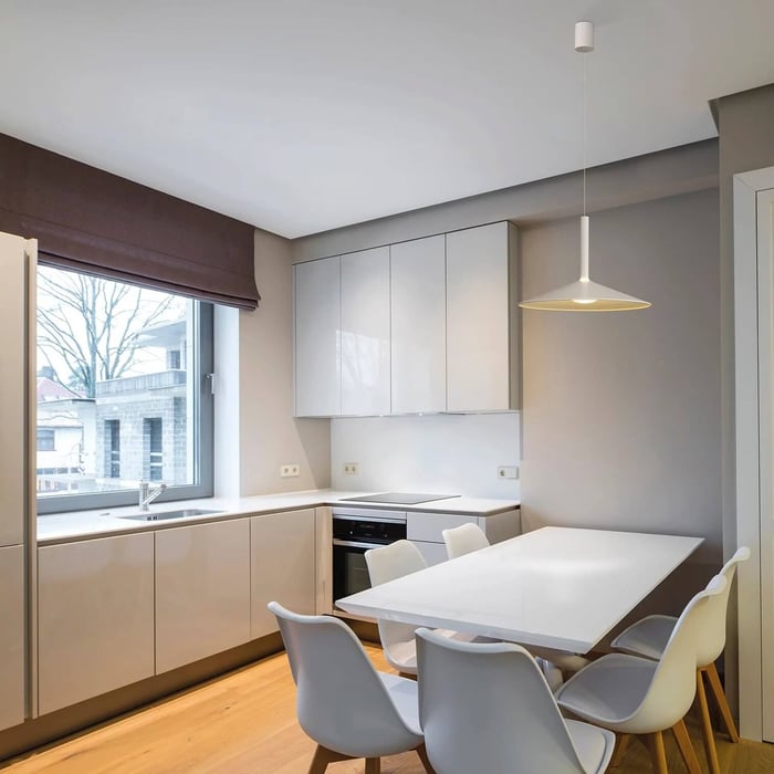 Compact light-grey kitchen dining nook with white cabinets, a windowed corner sink, and a single slim white pendant above a rectangular table.