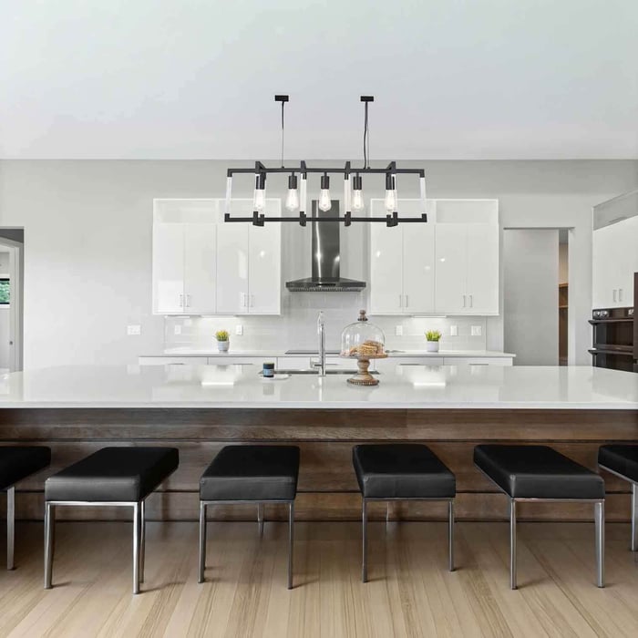 Black rectangular linear chandelier with exposed bulbs hanging above a white kitchen island with bar stools.