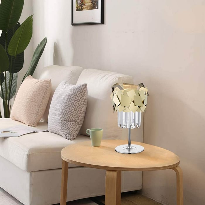Modern table lamp with geometric gold shade and crystal base on a round wooden side table in a neutral-toned living room.