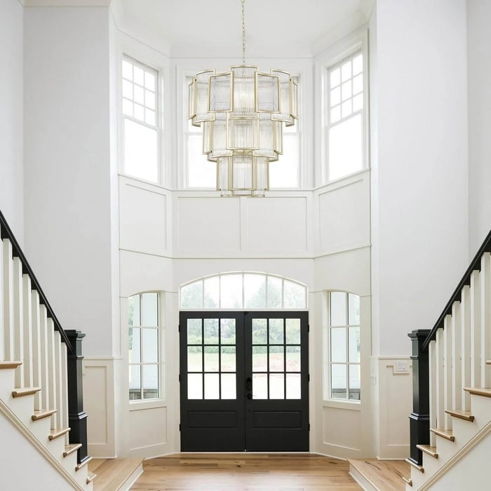 Tall entry hall with a tiered geometric chandelier hanging between double staircases and black framed doors.