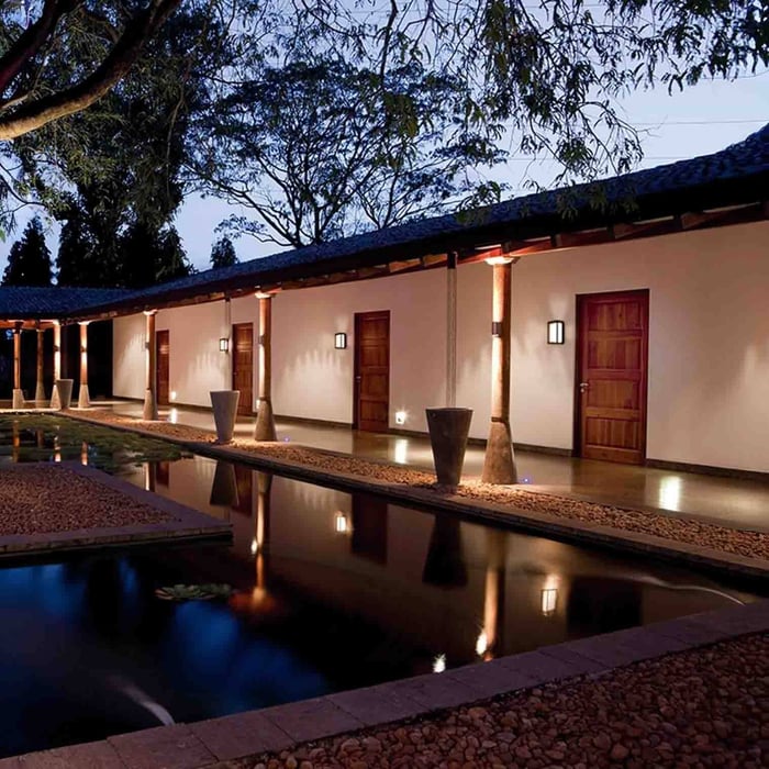 Long walkway beside a pool with warm wall lighting and uplights along a covered roofline.