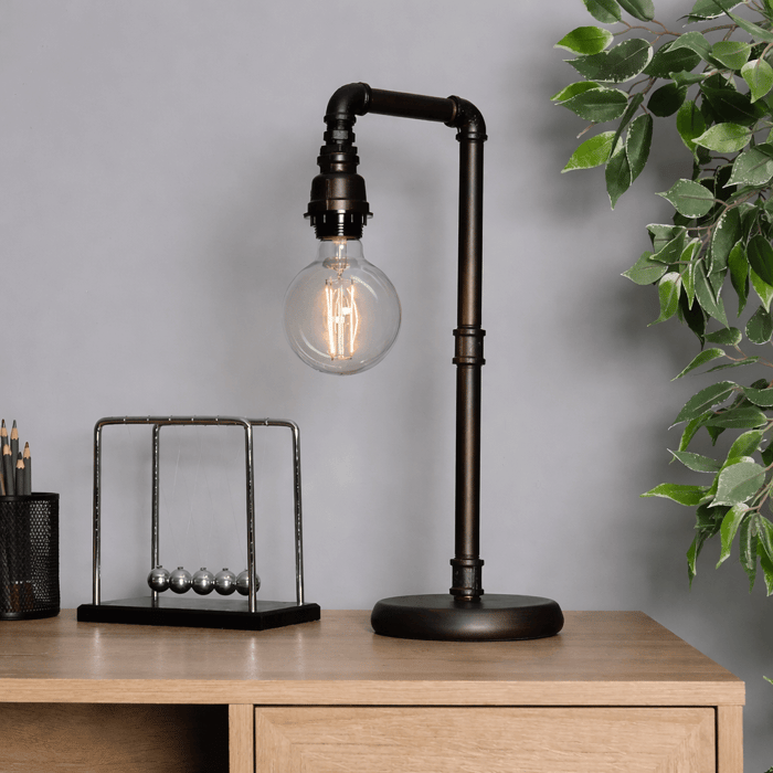Industrial table lamps with exposed filament bulb and dark metal pipe frame on a wooden desk, styled with a Newton’s cradle, pencil holder, and green plant.