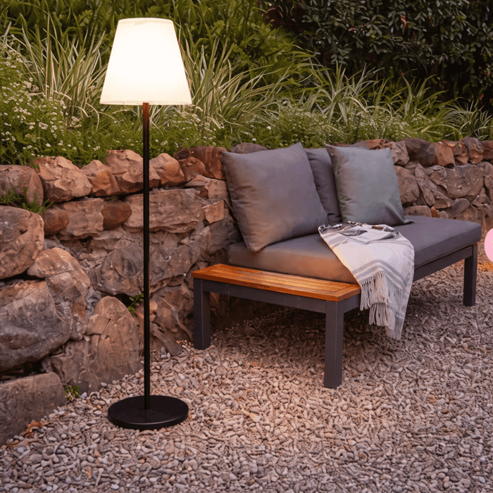 Slim black outdoor floor lamp with shade placed beside a cushioned daybed on gravel near a stone wall.