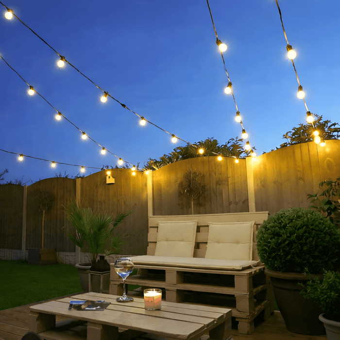 Outdoor string lights strung diagonally above a compact garden seating area with a pallet bench, coffee table, and candlelight at dusk.