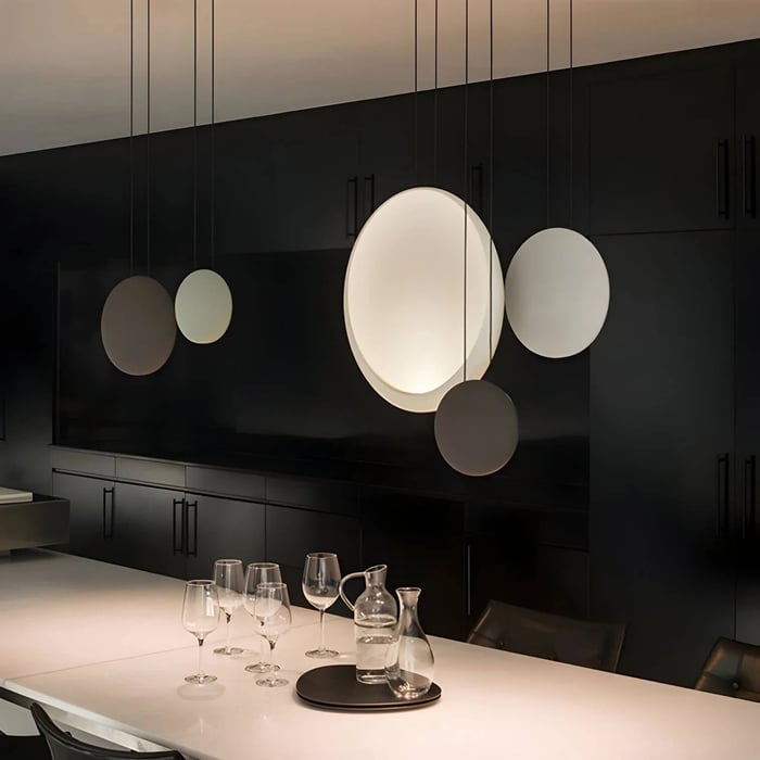 Moody black kitchen with a cluster of round disc pendant lights in different sizes above a white countertop set with glassware.