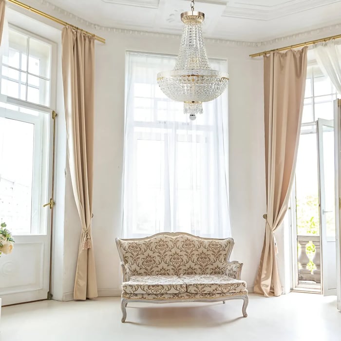 Bright living room with chandelier from the crystal chandeliers collection, vintage floral sofa, and tall beige curtains framing classic French doors.