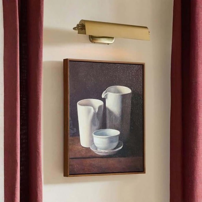 Brass picture light shining onto a framed still-life artwork on a pale wall, framed by deep red curtains.
