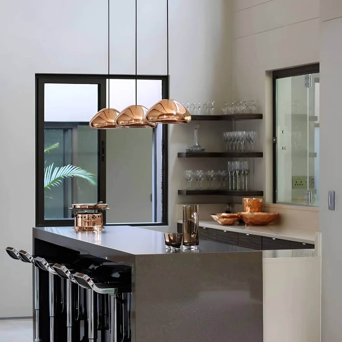 Modern kitchen with three copper designer pendant lights hanging above a sleek island with chrome bar stools and open shelves of glassware.