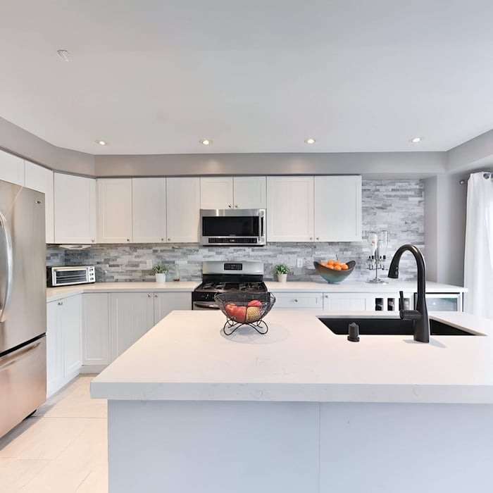 Bright kitchen with a large white island, grey stone backsplash, recessed ceiling downlights, and a black pull-out tap at the sink.