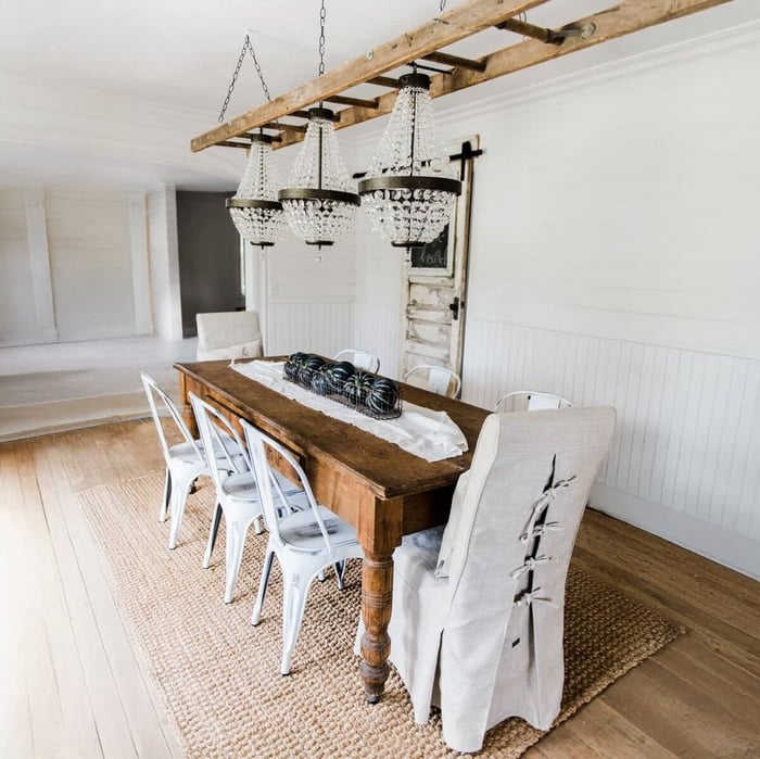Wooden table with white chairs under three rustic crystal chandeliers in a white-paneled dining room.