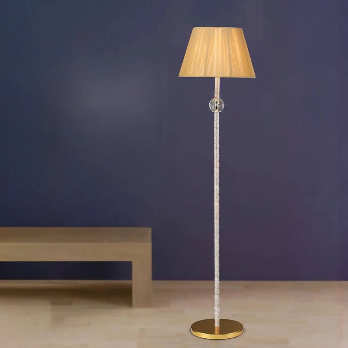 Tall crystal-effect floor lamp with gold base and beige pleated shade against a navy wall.