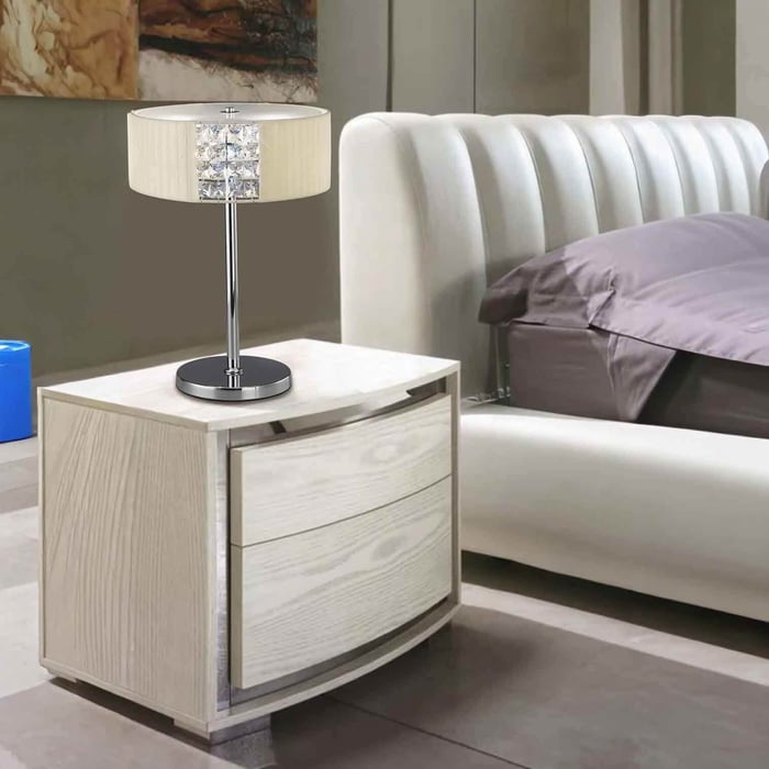 Chrome table lamp with a cream drum shade and crystal detail on a light wood bedside table next to an upholstered bed.