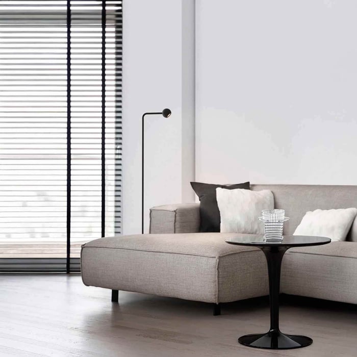 Minimal black floor reading lamp behind a grey sectional sofa near large window blinds.