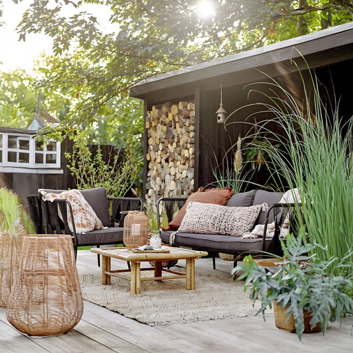 Wicker lounge furniture on a patio with natural lanterns and patterned cushions, surrounded by tall grass and stacked firewood.