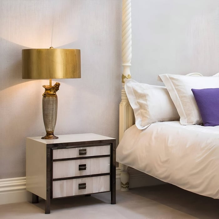 Bedroom with gold and ceramic bedside lamp, white bedding, and purple accent pillow.