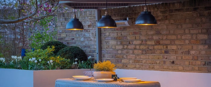 Brick-walled patio with a trio of industrial black dome pendant lights above an outdoor dining table set for two.