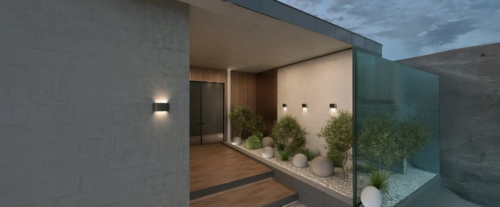 Modern home exterior with outdoor wall lights and minimalist garden featuring white stones and green shrubs at dusk.
