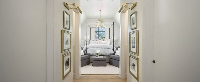 A luxurious bedroom with a modern canopy bed, plush grey armchairs, and gold accents. Wall-mounted picture lights illuminate framed art on the white walls, complementing the central geometric chandelier.