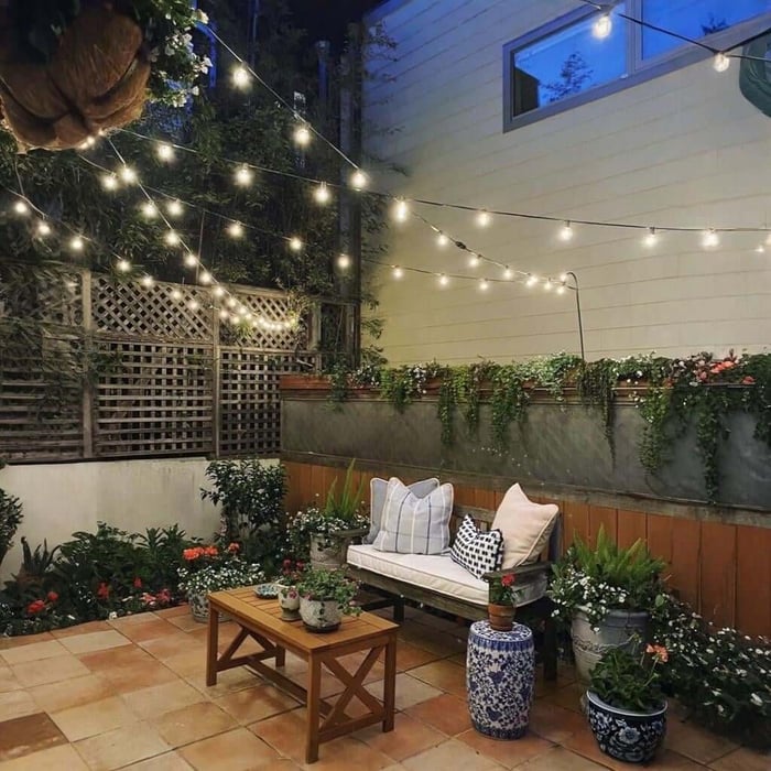 Outdoor string lights hung in layered strands over a small patio with a bench, coffee table, and potted flowers beside a trellis.