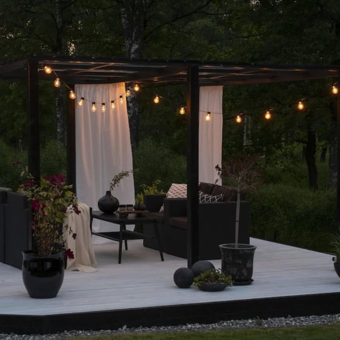 Outdoor string lights wrapped around a modern pergola with sheer curtains and dark outdoor furniture in a garden setting.
