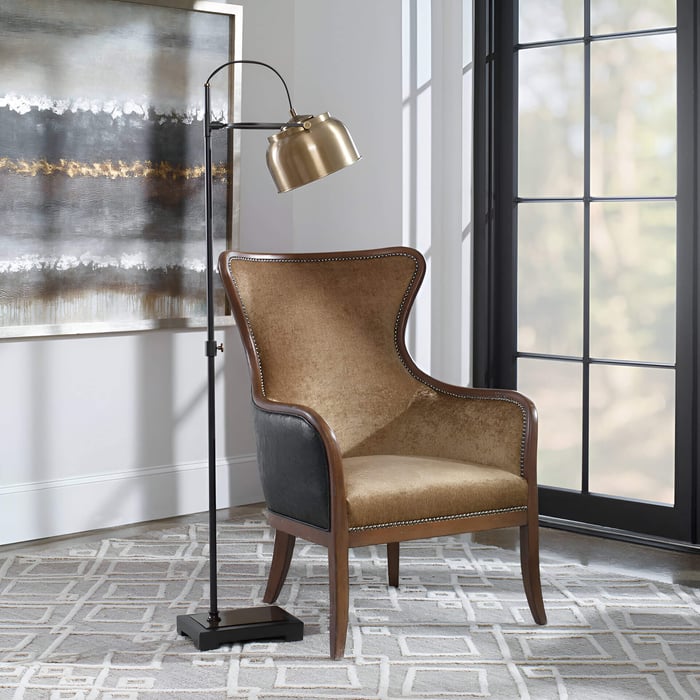 Brass-shaded floor lamp beside a tan upholstered accent chair in a bright living room with a large window and abstract wall art.