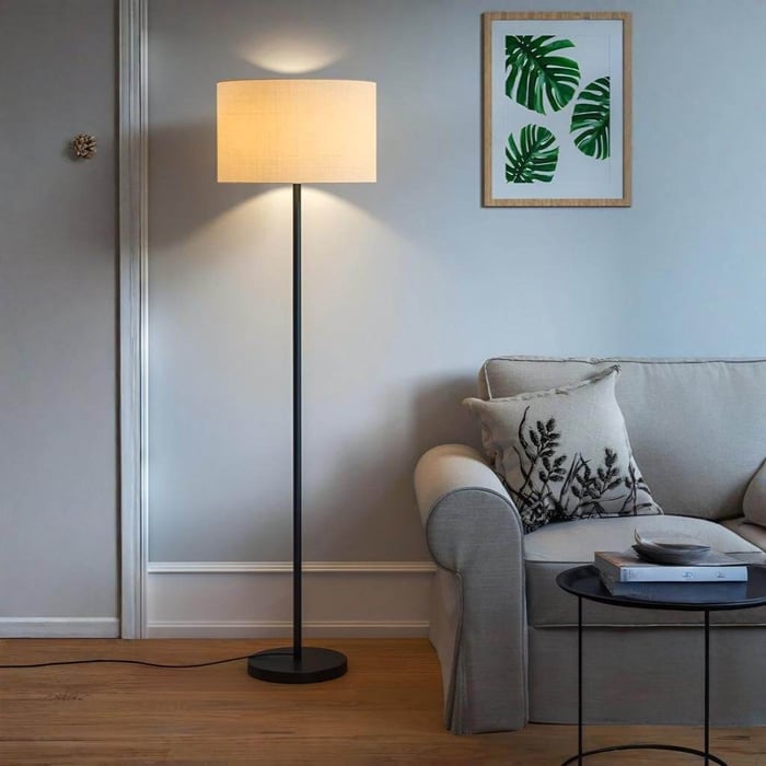 Black floor lamp with a beige drum shade illuminating a light gray sofa in a cozy living room with wooden floors and framed leaf artwork on the wall.