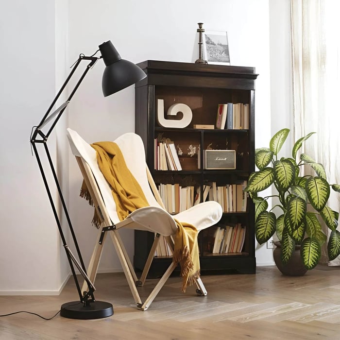 Black adjustable task floor lamp beside a white butterfly chair with a mustard throw in a cozy reading nook with a dark bookcase and large potted plant.