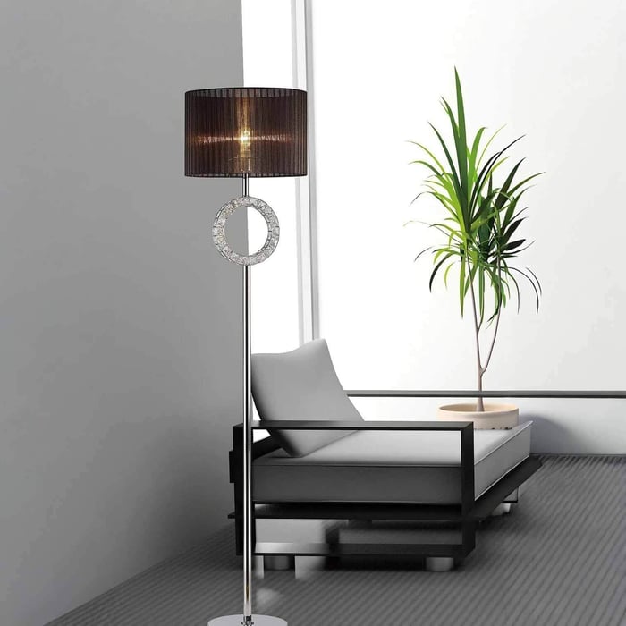 Chrome floor lamp with black drum shade and crystal ring accent beside a lounge chair and potted plant by a window.