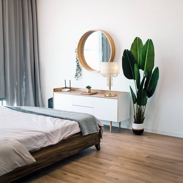 Bright bedroom corner with a wooden dresser, gold-accented table lamp, round wall mirror, and a large indoor plant.