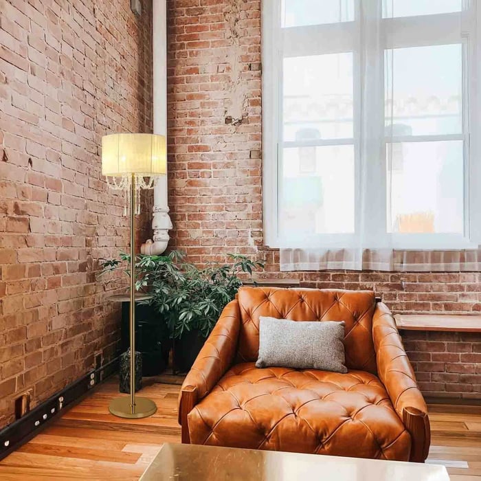 Gold floor lamp with pleated shade beside a tan tufted leather armchair in an exposed brick loft with large windows.