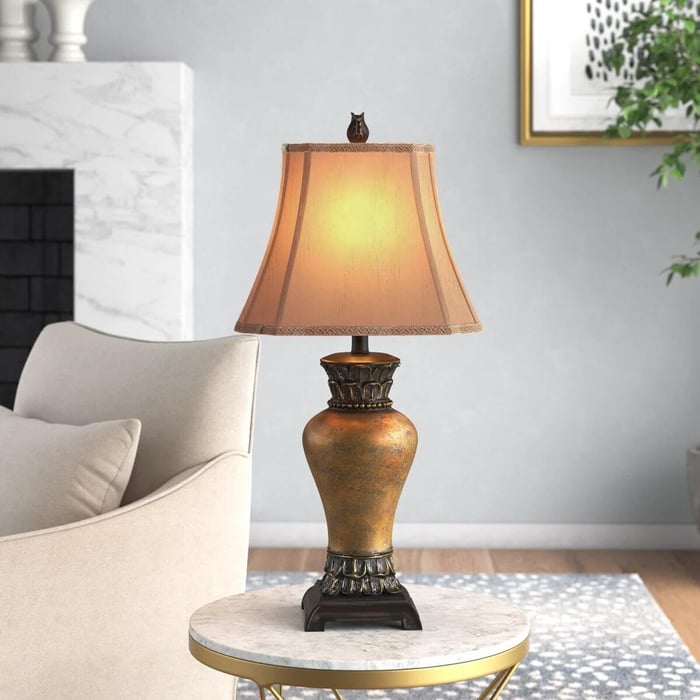 A traditional table lamp with a warm amber shade and ornate bronze base sits on a round marble-top side table next to a cream armchair in a cozy living room.