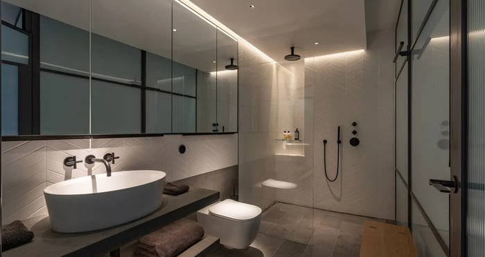 A sleek and contemporary bathroom with herringbone wall tiles and recessed ambient lighting. It includes a floating vanity with a vessel sink, a walk-in shower with matte black fixtures, and a minimalist aesthetic.
