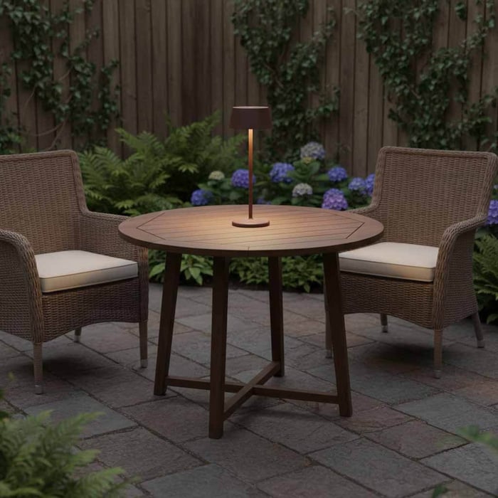 Round wooden patio table with two wicker chairs and a slim outdoor table lamp in the centre, set against garden plants and a timber fence.