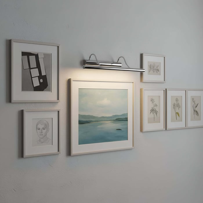 Gallery wall of framed artwork with a picture light above a central landscape print on a pale textured wall.